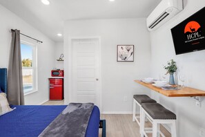 Ein Schlafzimmer mit einem Bett, einem roten Gerät, einem Fernseher, einem Fenster und einem Essbereich mit Tisch und Bänken.