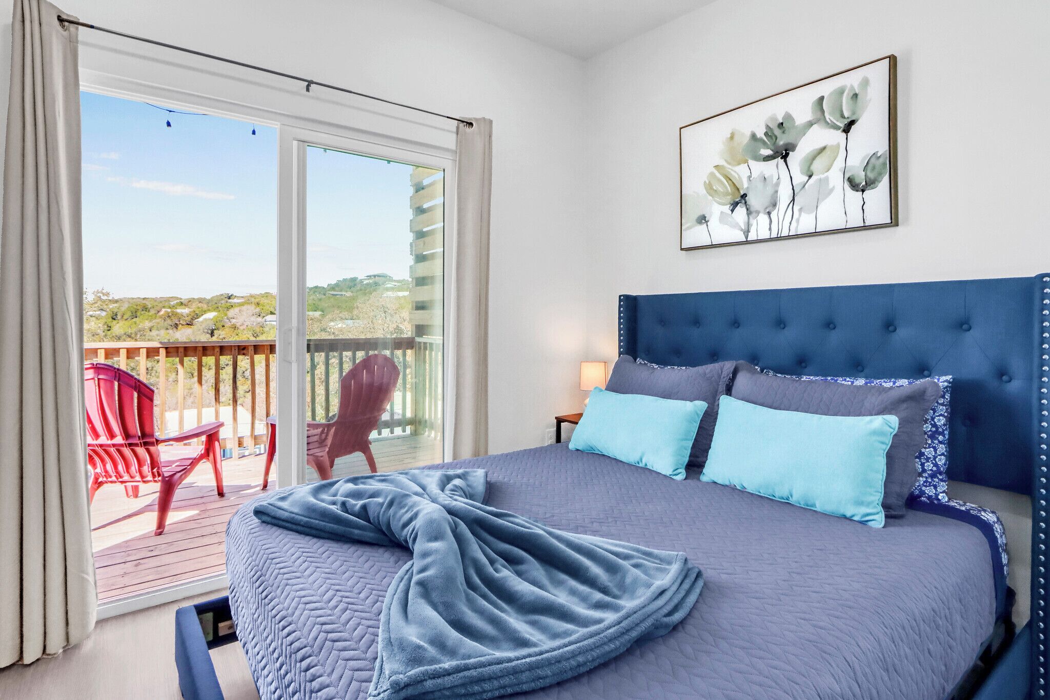A bedroom with a large bed, a blue headboard, a painting, and a balcony with red chairs.