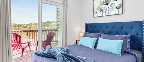 A bedroom with a large bed, a blue headboard, a painting, and a balcony with red chairs.