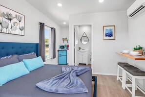 A bedroom with a bed, a bedside table, a desk, a chair, a window with curtains, a wall-mounted air conditioner, and a framed picture. - Hill Country Retreat w/ Canyon Lake Views (Canyon Lake)