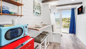 Una cocina con microondas, un electrodoméstico rojo, un estante de madera con objetos, una mesa con una silla y vistas a través de una puerta.