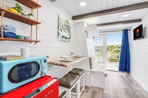 A kitchen with a microwave, a red appliance, a wooden shelf with items, a table with a chair, and a view through a door. - Modern Container Escape Near Canyon Lake (Canyon Lake)