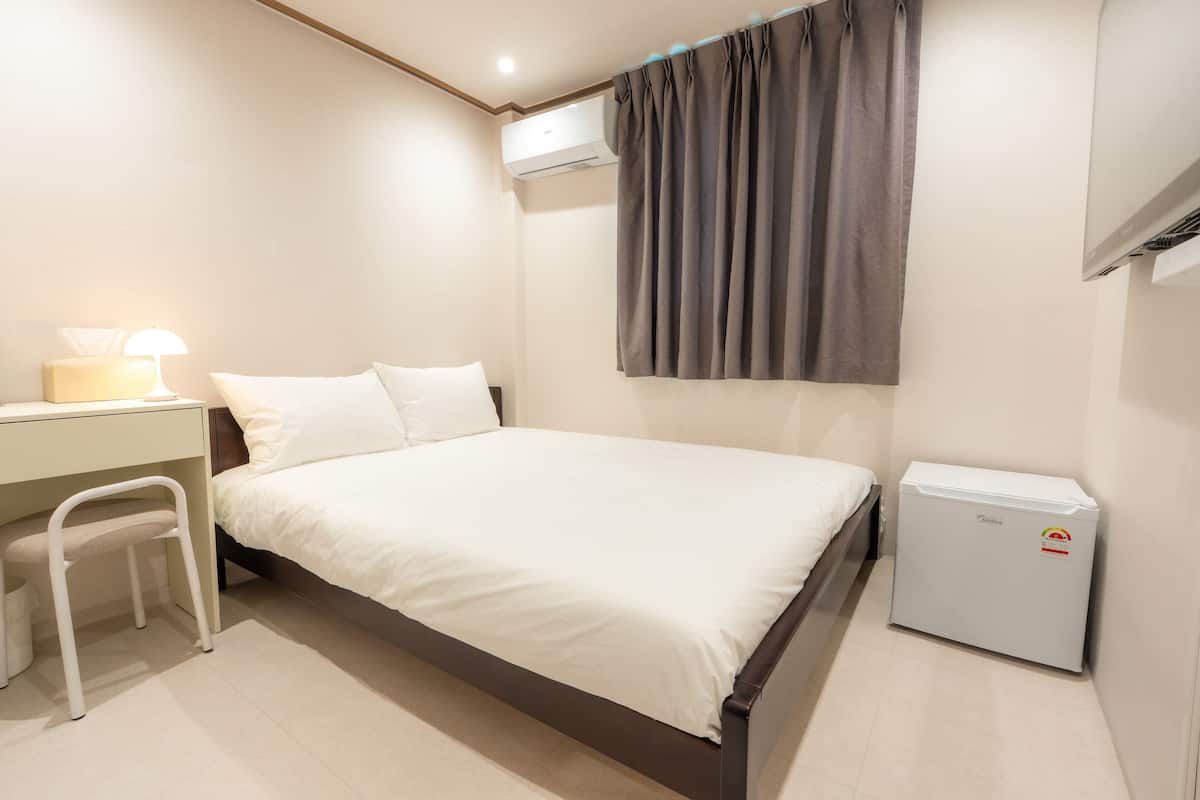 A bedroom with a bed, a desk, a chair, an air conditioner, and a mini fridge.