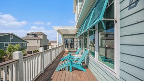 A wooden deck with chairs and a covered area, overlooking a residential neighborhood. - Beachside Vibes | Luxury Gulf Front Home (Galveston)