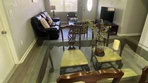 Un comedor con una mesa de vidrio y sillas, un sofá, un televisor y una ventana con persianas.
