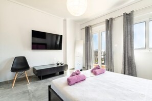 A modern hotel room with a large bed, a flat-screen TV, a small desk, and a balcony with curtains. - Evergreen Loft | Modern City Centre Living (Kos)