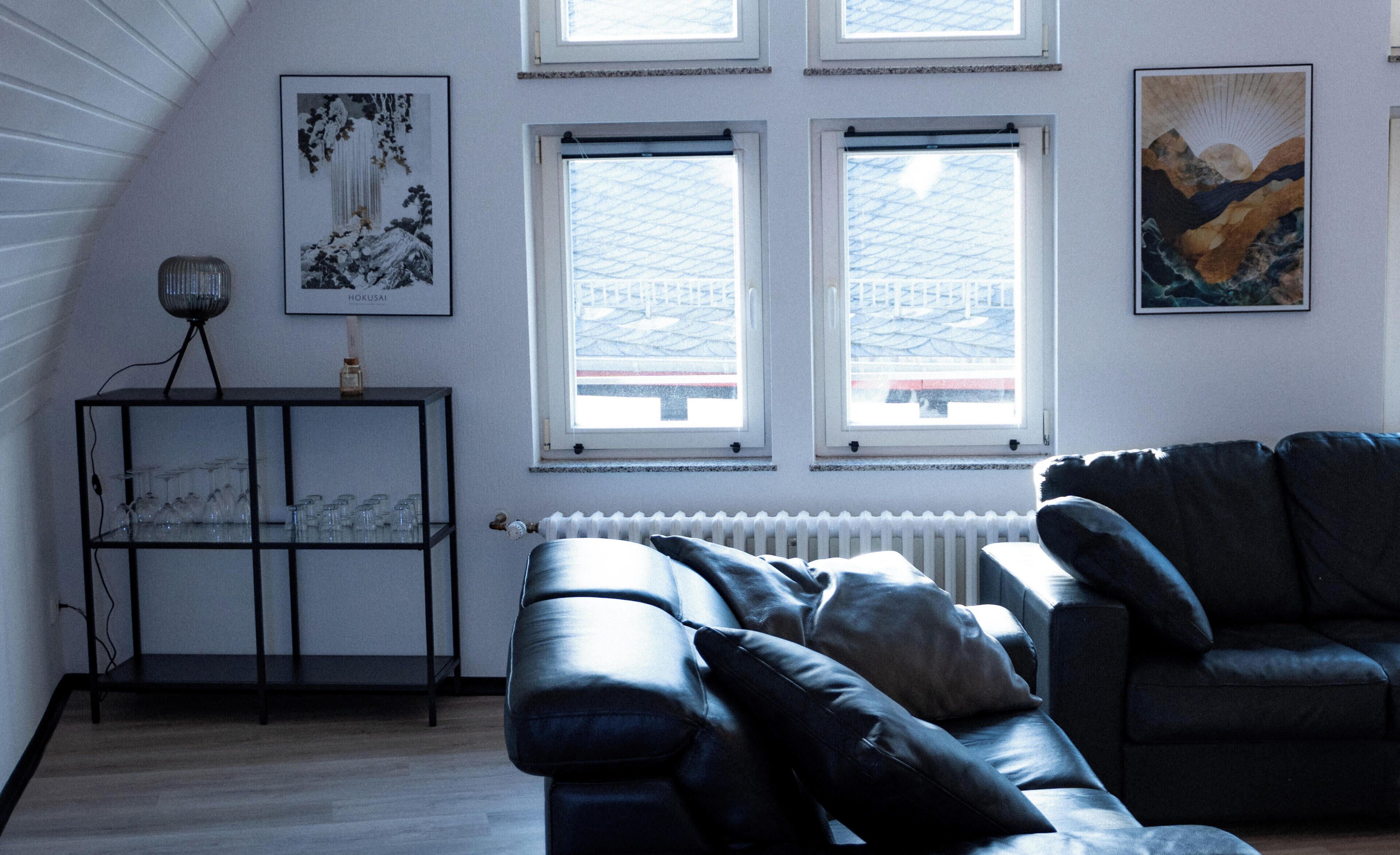 A living room with a black leather sofa, a glass cabinet with wine glasses, and two windows with a view of a snowy landscape.