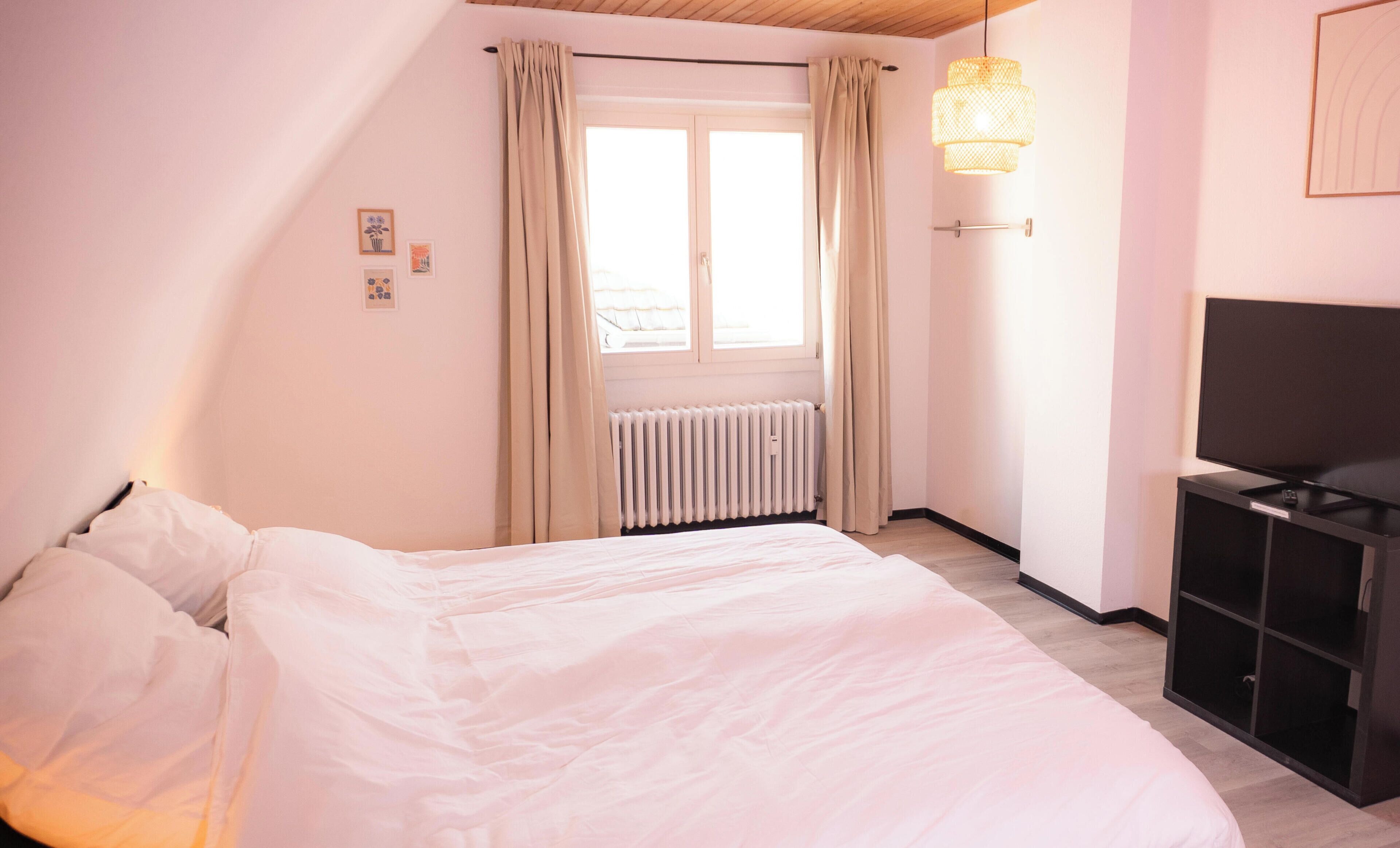 Een slaapkamer met een bed, een radiator, een televisie en een raam met gordijnen.
