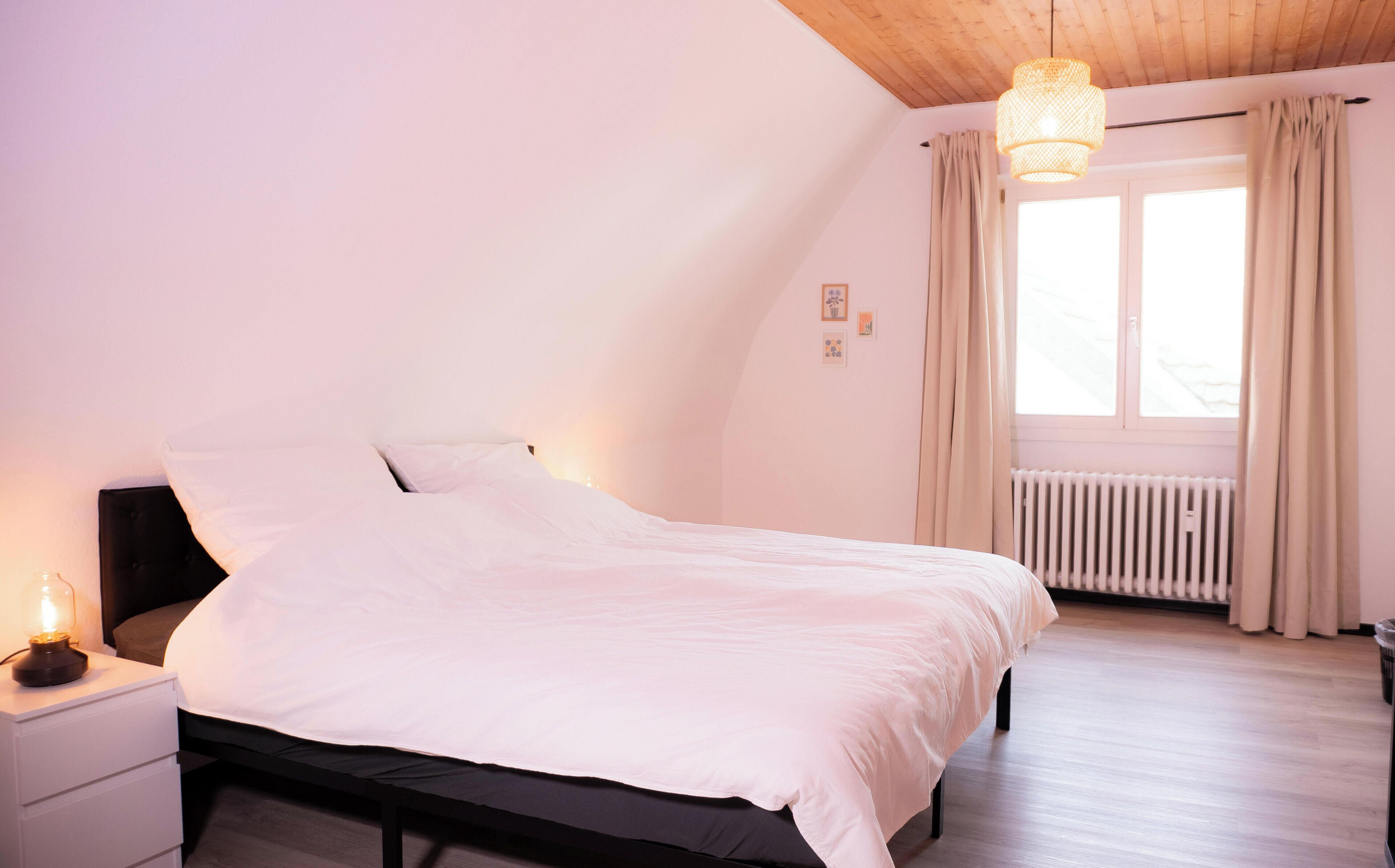 Un dormitorio con cama, lámpara de noche, ventana con cortinas y un radiador.