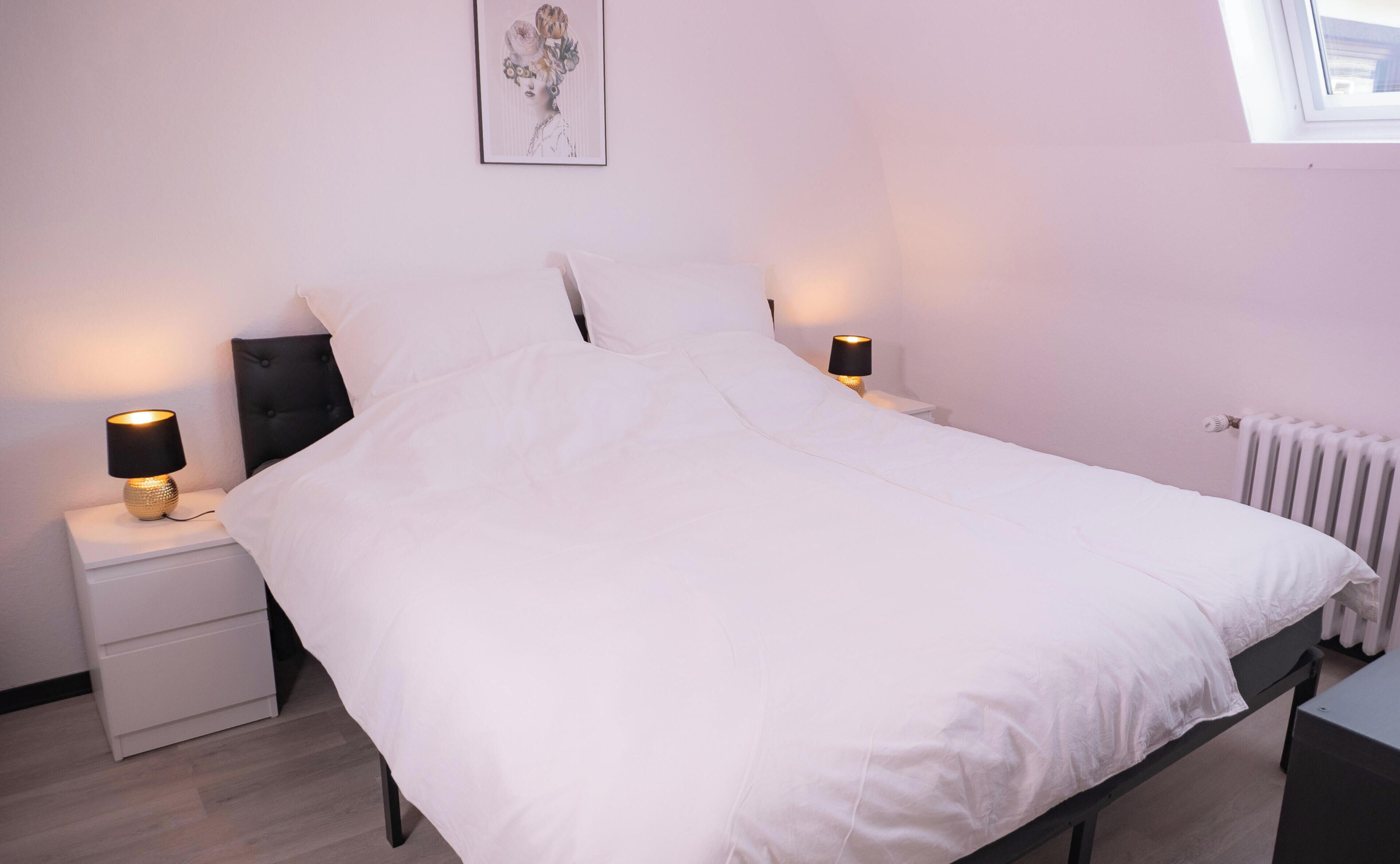 Une chambre à coucher comprenant un lit, deux tables de chevet, une commode, un radiateur et une fenêtre.