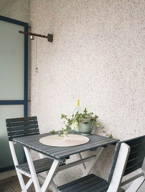 Ein kleiner Tisch im Freien mit einer gewebten Tischmatte, umgeben von zwei Stühlen, vor einer strukturierten Wand mit blauer Umrandung.
