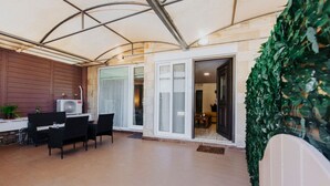 Un patio cubierto con mesa y sillas, un ventilador grande y vistas a la habitación contigua.
