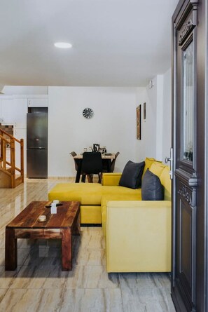 Ein Wohnzimmer mit gelber Couch, einem Holztisch, einem Essbereich mit Tisch und Stühlen sowie einer Küche mit Kühlschrank.