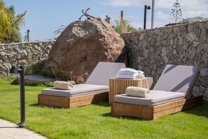 Twee houten lounge stoelen met kussens en handdoeken, geplaatst tegen een stenen muur en een grote rots, op een gazon.