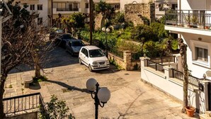 A residential area with a white car parked on a paved street, surrounded by buildings with balconies and a palm tree.