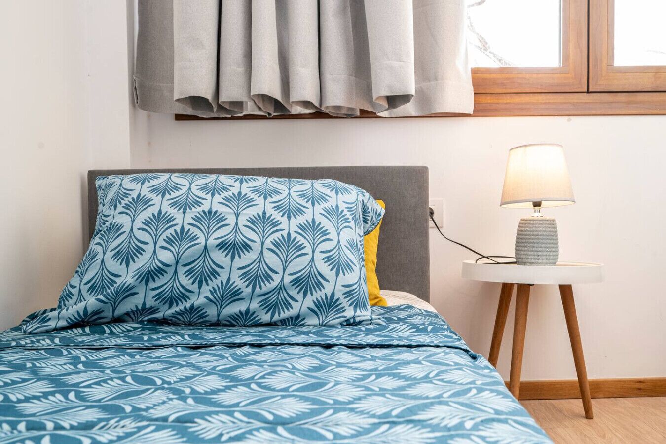 Uma cama com colcha azul e branca estampada, cabeceira cinza e travesseiro amarelo. Há uma mesa de cabeceira com abajur e base branca.