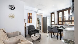Uma sala de estar moderna com um sofá, uma mesa redonda pequena e uma poltrona. Há uma área de refeições com uma mesa e cadeiras, e uma cozinha com uma geladeira e uma janela.