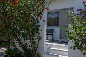 A white building with a door and window, a black chair outside, and a tree with red flowers. - Thines Studios : Studio A (Kampos)