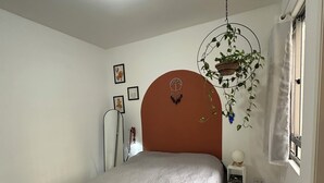 Un dormitorio con cama, luz de techo, planta en la pared, ventana con cortinas y mesita de noche con lámpara.