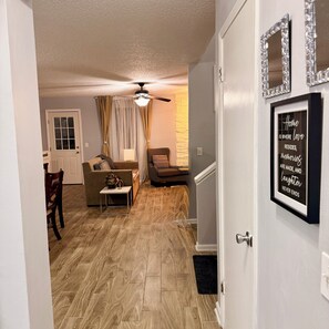 Un salon doté d’un ventilateur de plafond, d’un canapé, d’une table basse et d’une porte ornée d’un miroir et d’une citation encadrée.