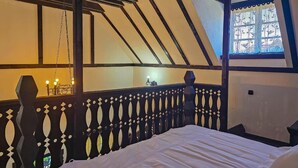 Quarto com teto inclinado, uma cama, janela com cortina decorativa e uma pequena mesa com abajur.