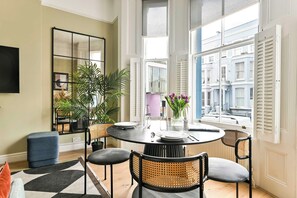 A dining area with a round table, four chairs, a potted plant, and a view of buildings through large windows. - The Notting Hill Blue Palace (Greater London)