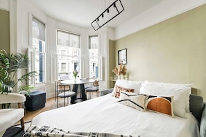 A hotel room with a bed, a round dining table, chairs, a potted plant, and a view of buildings through large windows. - The Notting Hill Blue Palace (Greater London)
