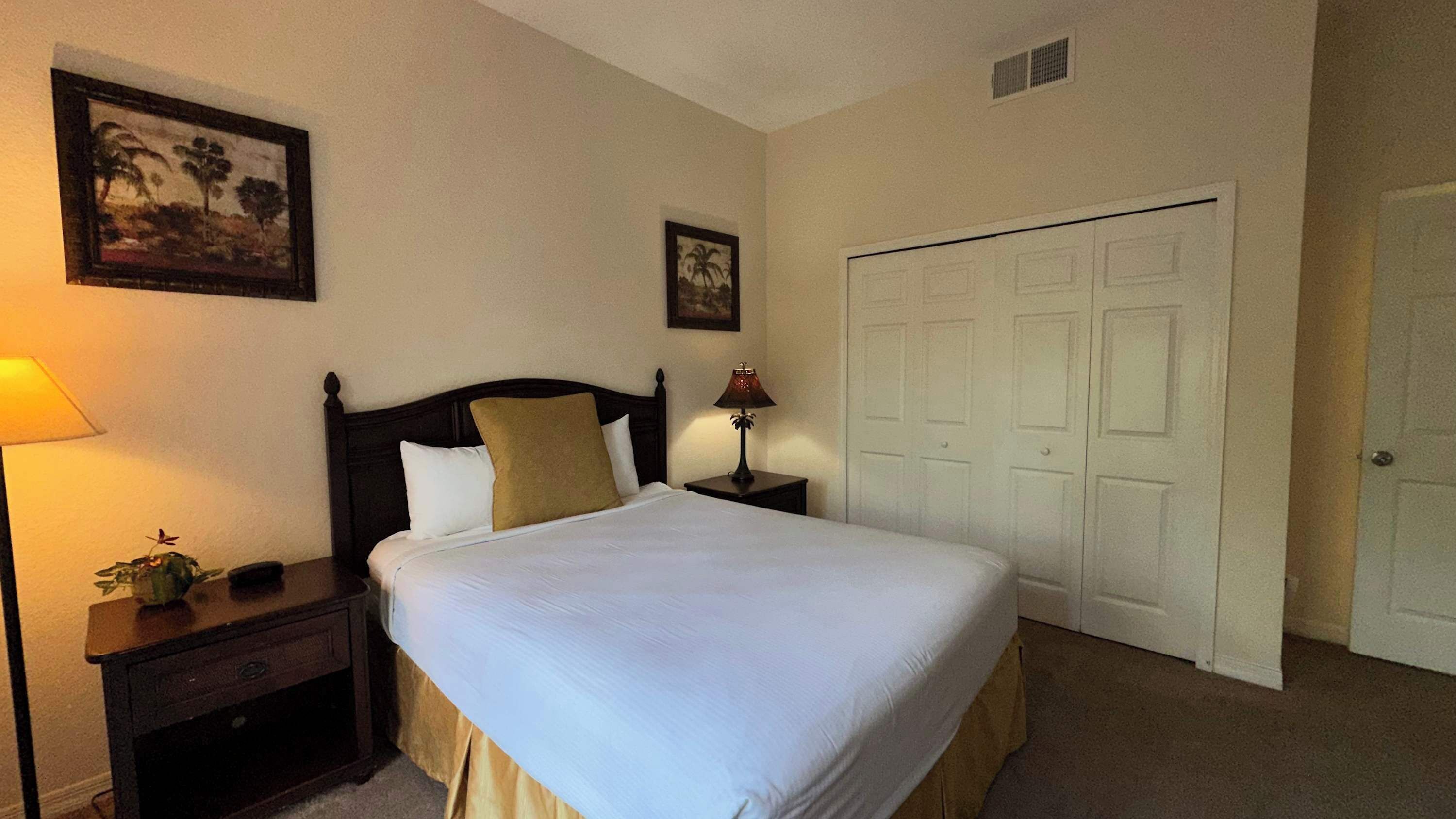 A neatly made bed with white linens, a dark wooden headboard, and two bedside lamps. There are two framed pictures on the wall, a door with a handle, and a small plant on the nightstand.
