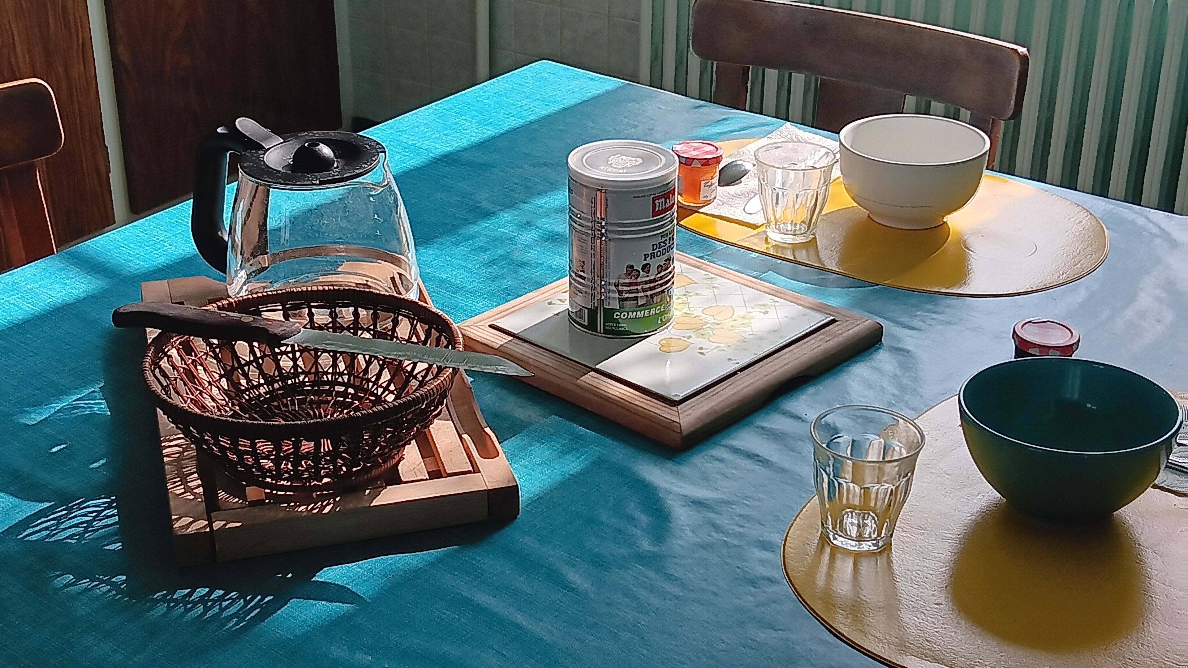 A dining table with a blue tablecloth, a basket, a knife, a glass pitcher, a can, a glass, and a bowl.