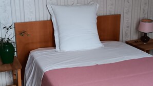 A neatly made bed with a white pillow and a pink blanket, a green plant on a wooden nightstand, and a pink lamp on a wooden nightstand.