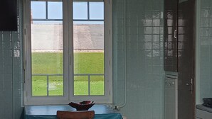 A dining area with a table set for two, a window with a view of a grassy field, and a kitchen area visible through an open door.