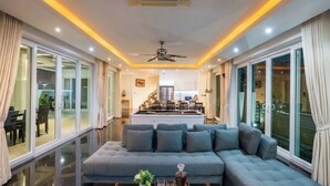 A modern living room with a large grey sectional sofa, a ceiling fan, and a kitchen in the background. - Luxury 5BR Villa Jomtien - Pool, Sports & Karaoke Close to the beach of pattaya (เมืองพัทยา)