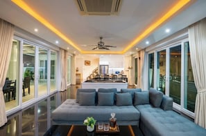 A modern living room with a large grey sectional sofa, a ceiling fan, and a kitchen in the background. - Luxury 5BR Villa Jomtien - Pool, Sports & Karaoke Close to the beach of pattaya (เมืองพัทยา)
