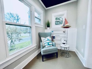 Habitación de esquina con ventana, una silla, una mesita y una estantería con una planta y un barco modelo.