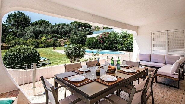 Un espace extérieur abrité, aménagé pour les repas, avec une table pour quatre personnes, donnant sur une piscine et un jardin.