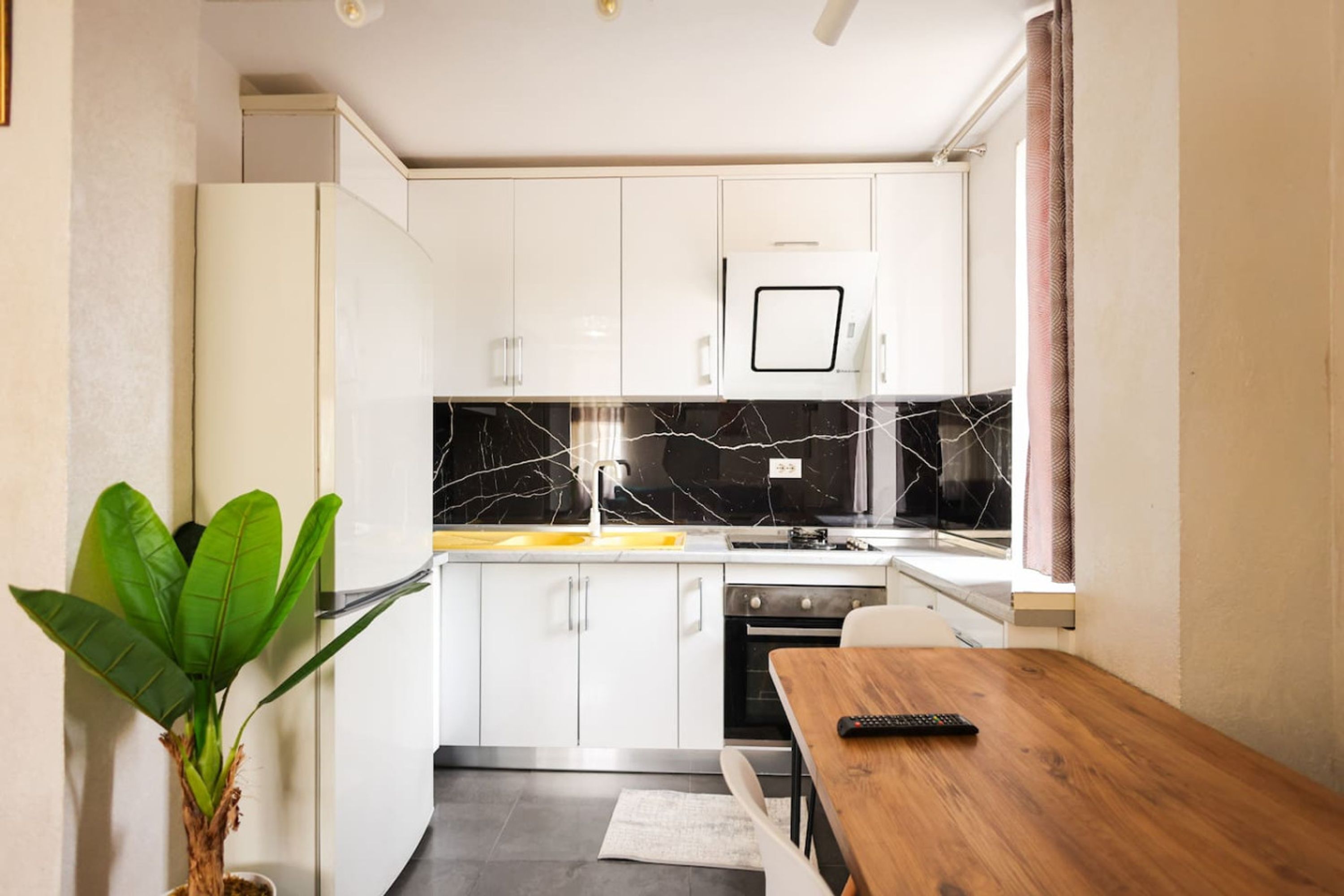 Una cucina compatta con mobili bianchi, una parete di fondo in marmo, un tavolo da pranzo in legno e una pianta in vaso.