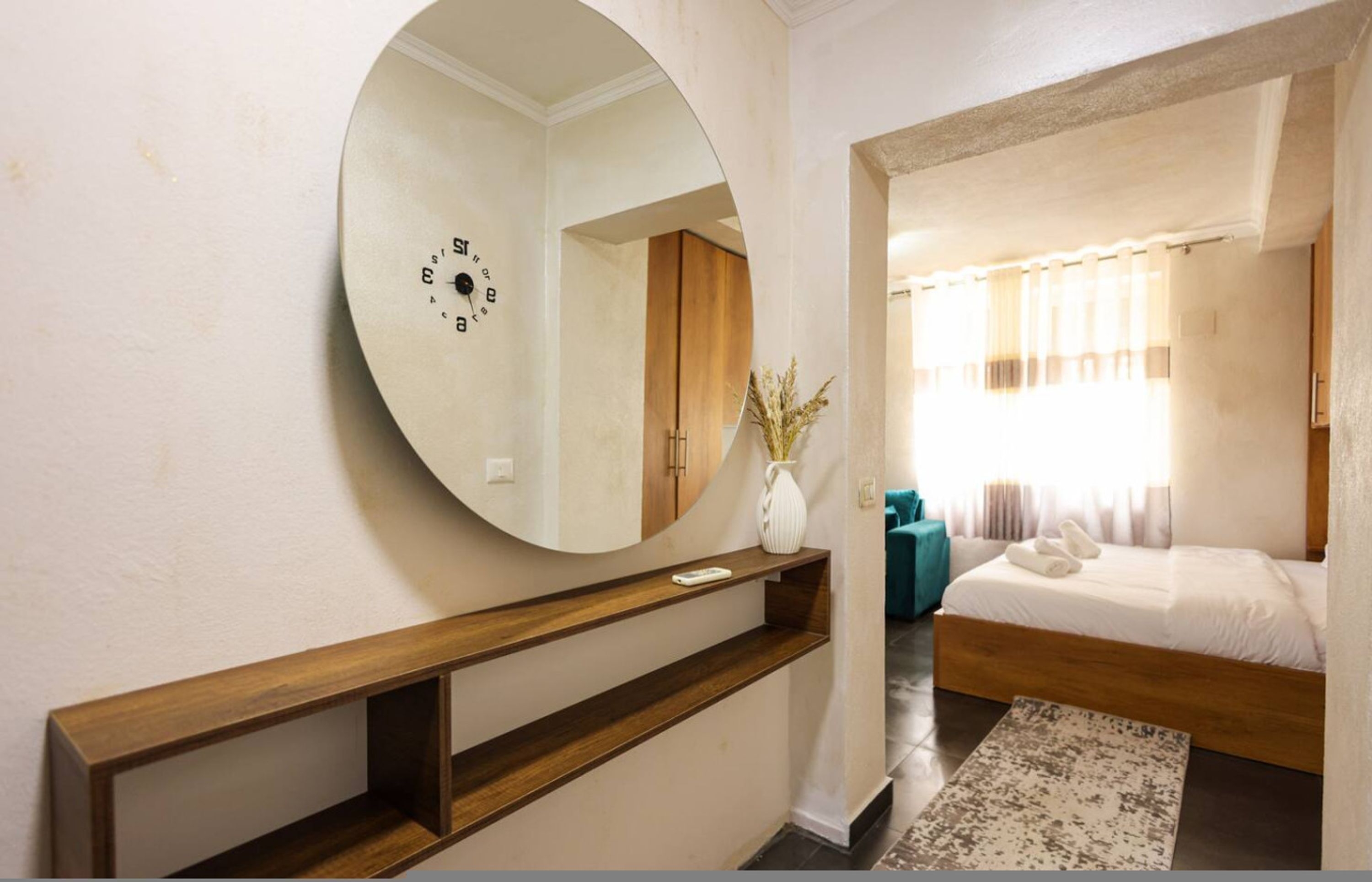 Een slaapkamer met een houten bed, een ronde spiegel, een kleine witte vaas en een raam met gordijnen.
