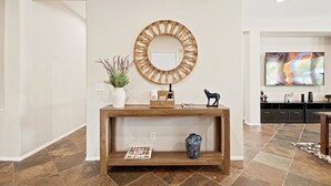 Een woonkamer met een houten console, een ronde spiegel, een plant in pot, een vaas en een televisie aan de muur.