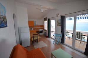 Ein Zimmer mit Esstisch, Stühlen, einer Couch, einem kleinen Tisch, Deckenventilator, einer Küchenzeile und einer Glastür, die auf einen Balkon mit Meerblick führt.