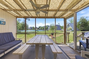 Une terrasse couverte avec une table et des bancs en bois, un ventilateur de plafond et une vue sur un lac et une zone herbeuse.