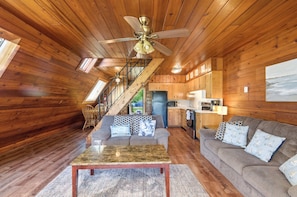 LâintĂ©rieur dâun chalet en bois comprend un escalier, un salon avec un canapĂ© et une table basse, ainsi quâun coin cuisine.