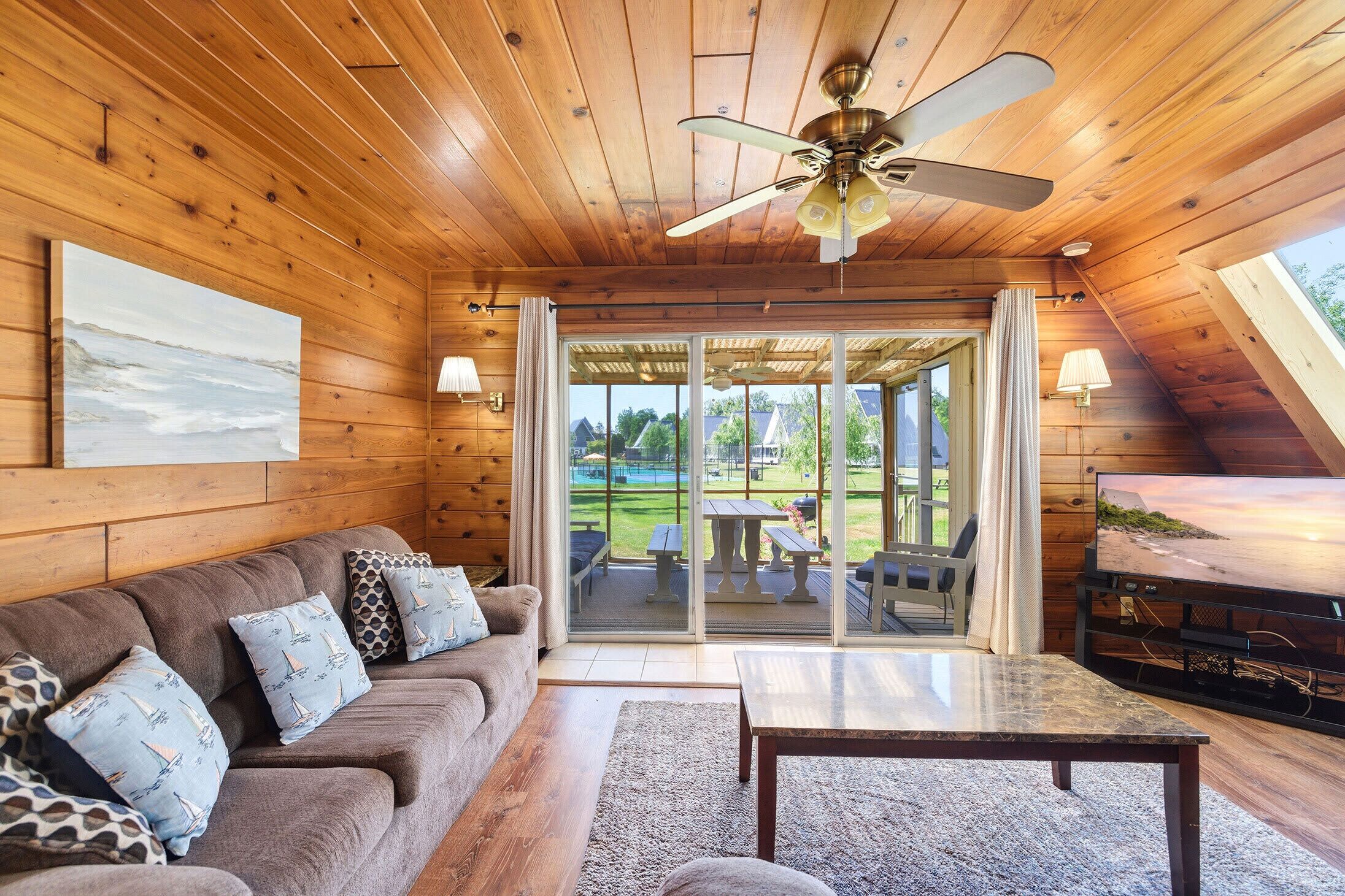 Ein gemütliches Zimmer mit Holzbalkendecke, Deckenventilator, einer Couch mit gemusterten Kissen, einem Tisch und einem Fernseher.