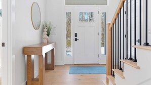 A hallway with a wooden console table, a mirror, a door with a window, and a staircase with black railings. - Van Duyne North Airy Escape (Ogunquit)