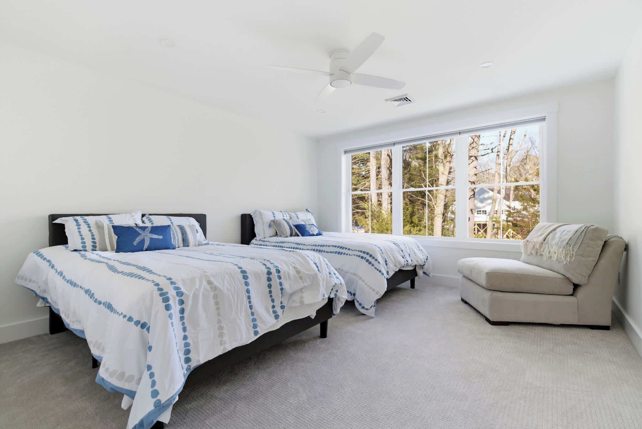 A bedroom with two beds, a ceiling fan, a large window with a view of trees, and a beige armchair.