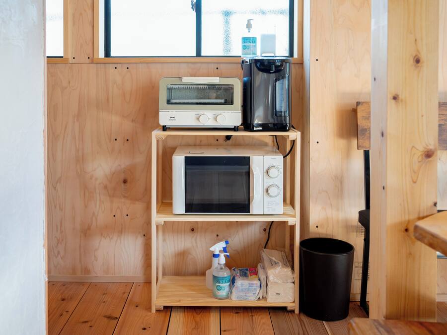 Une petite cuisine équipée d’un micro-ondes, d’un grille-pain et d’une machine à café, le tout posé sur une étagère en bois, ainsi qu’une poubelle et des produits de nettoyage sur le sol.