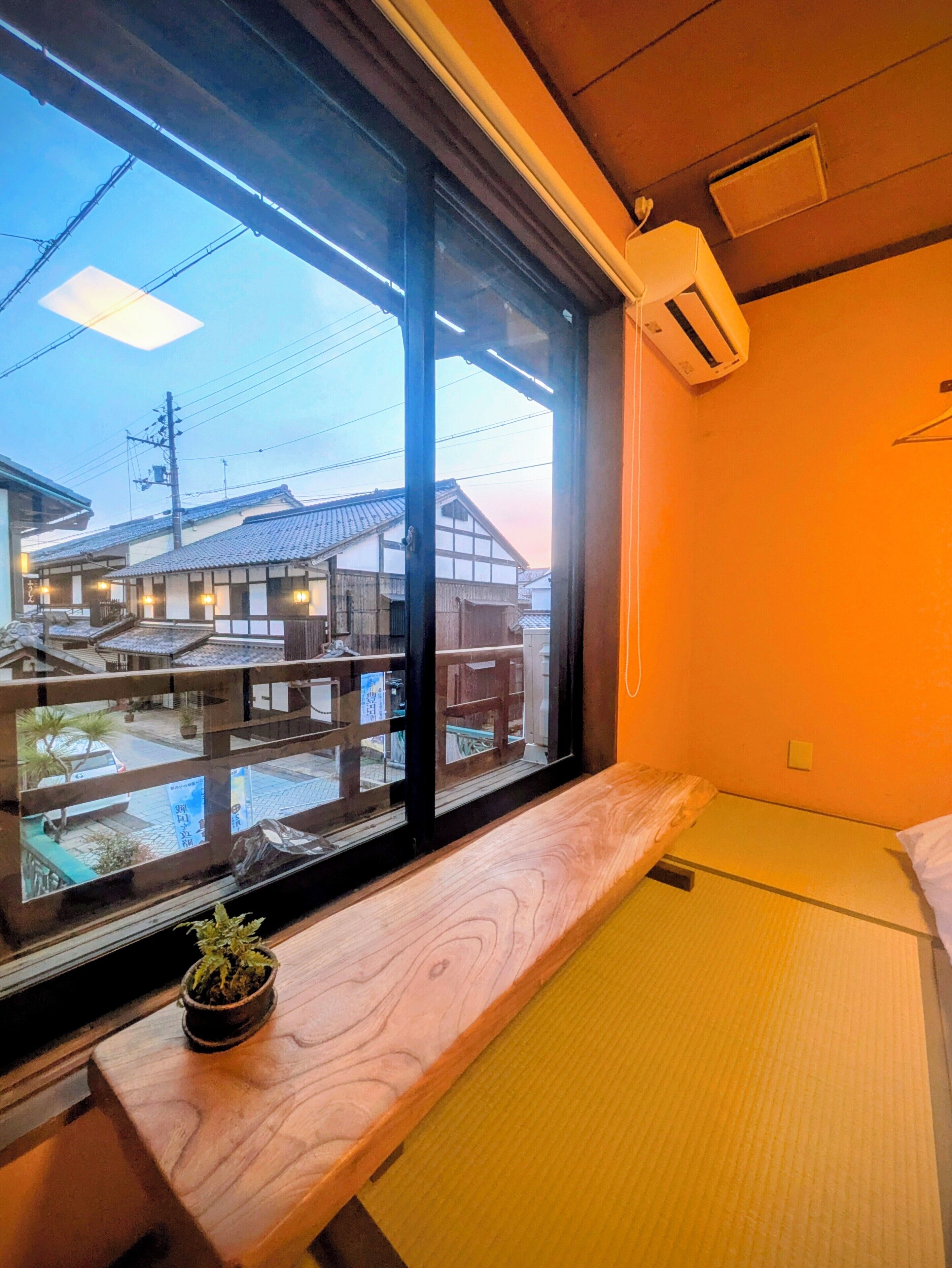 A room with a large window, a wooden bench, a small potted plant, and a bed with a futon.