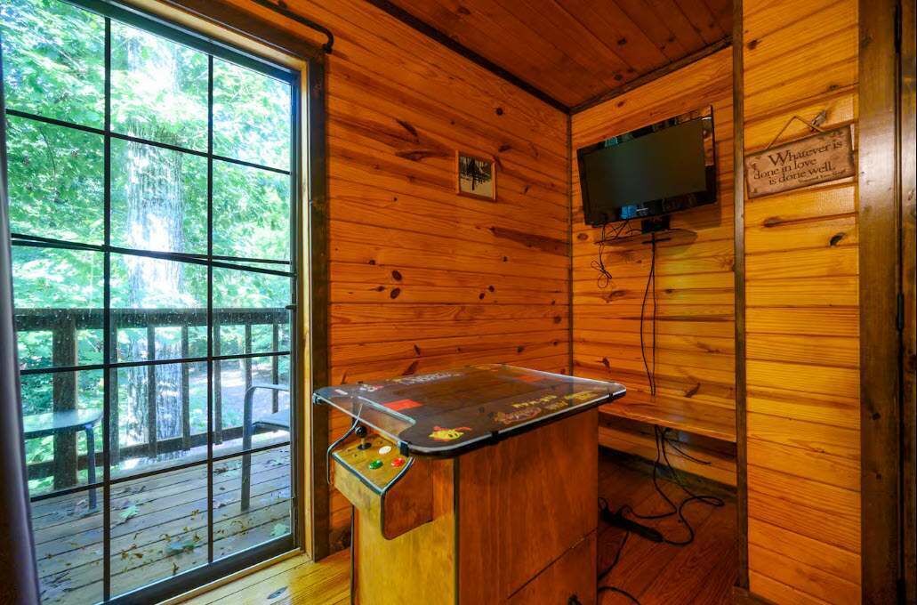 Interior de uma cabana de madeira com mesa de madeira, televisão na parede e janela com vista para árvores.