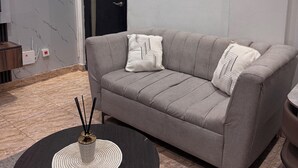 Una sala de estar moderna con un sofá gris, dos almohadones decorativos, una mesa de centro negra redonda con un posavasos tejido, un cuadro abstracto colgado en la pared y una puerta con pomo negro.