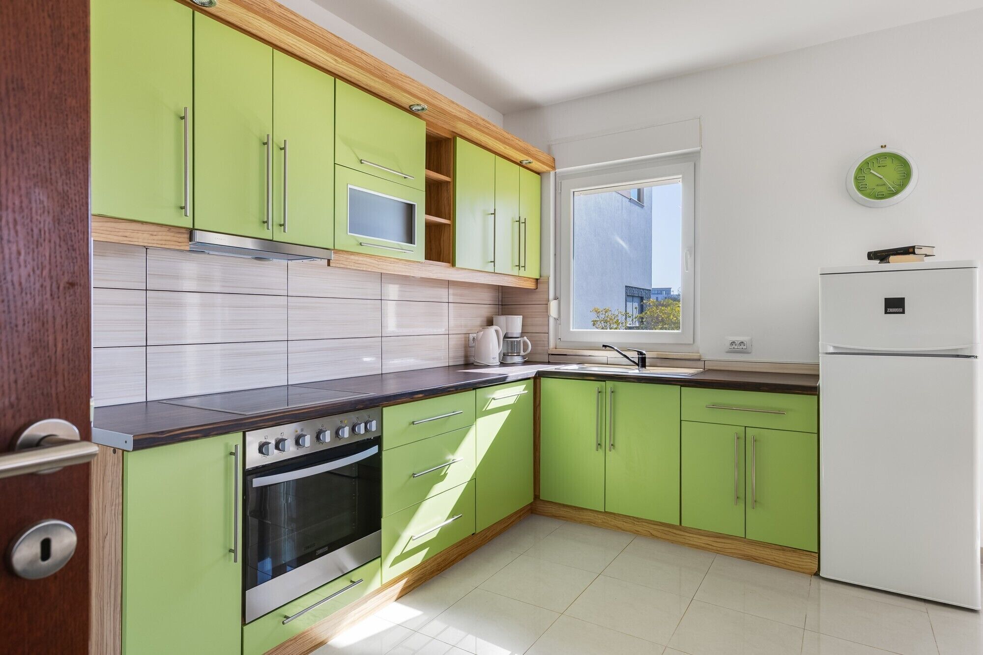 Une cuisine avec des meubles de couleur verte, un four encastré, un réfrigérateur blanc et une fenêtre donnant sur un beau paysage.
