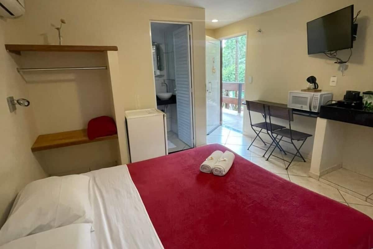A bedroom with a bed, a red blanket, a small kitchenette, a TV, and a door leading to a balcony.